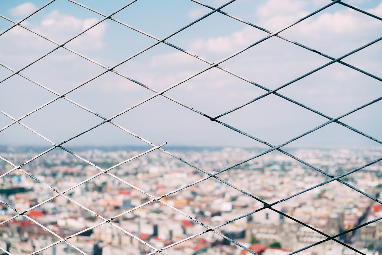 Blurred cityscape view through a metal chain link fence, showcasing depth and urban pattern.