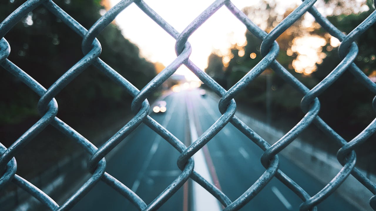 A blurred view of a highway through a chain-link fence at sunset, capturing the essence of urban transit.
